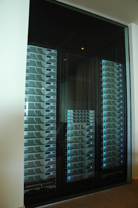 A server room or data center featuring several racks of network servers with blue indicator lights. The setup is organized and enclosed within a glass cabinet.