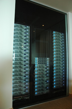A server room or data center featuring several racks of network servers with blue indicator lights. The setup is organized and enclosed within a glass cabinet.