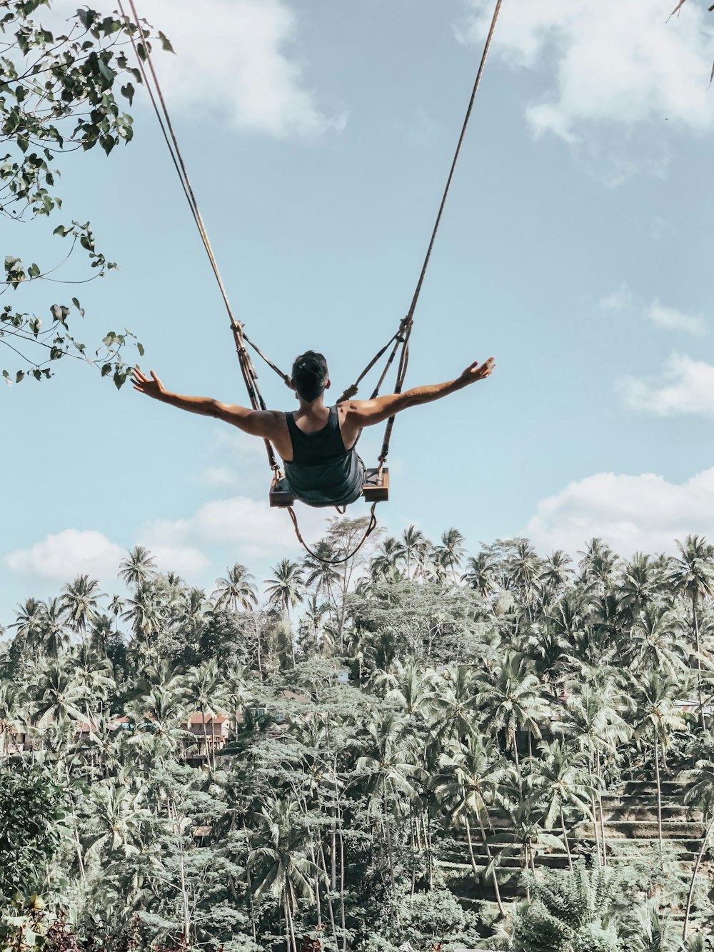 man wearing black tank top sitting on outdoor swing while raising both hands viewing trees under blue and white sky