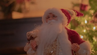 An image of a festive holiday scene featuring a Santa Claus figure with a white beard and red hat. The Santa figure appears to hold a plush toy and is set against a background blurred with warm light and holiday decorations.