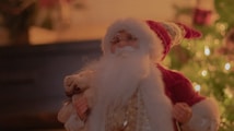 An image of a festive holiday scene featuring a Santa Claus figure with a white beard and red hat. The Santa figure appears to hold a plush toy and is set against a background blurred with warm light and holiday decorations.