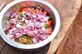 A large metal bowl contains a mix of chopped red onions, tomatoes, cilantro, and possibly other raw vegetables or herbs. The bowl is placed on a wooden surface, and the image suggests a fresh and rustic setting.