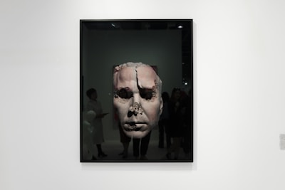 A framed artwork features a large, textured face that appears to be eroded or damaged. The piece is displayed in a gallery setting with people observing it. The gallery is well-lit, and the artwork contrasts against the plain white wall.