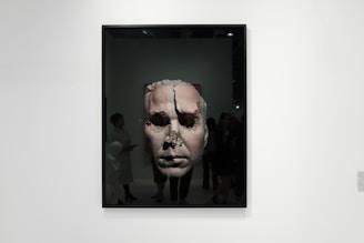 A framed artwork features a large, textured face that appears to be eroded or damaged. The piece is displayed in a gallery setting with people observing it. The gallery is well-lit, and the artwork contrasts against the plain white wall.