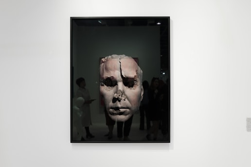 A framed artwork features a large, textured face that appears to be eroded or damaged. The piece is displayed in a gallery setting with people observing it. The gallery is well-lit, and the artwork contrasts against the plain white wall.