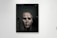 A framed artwork features a large, textured face that appears to be eroded or damaged. The piece is displayed in a gallery setting with people observing it. The gallery is well-lit, and the artwork contrasts against the plain white wall.