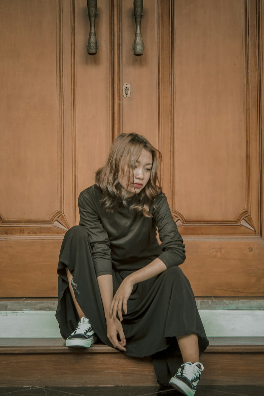 woman wearing black crew-neck long-sleeved dress sitting near closed brown wooden door