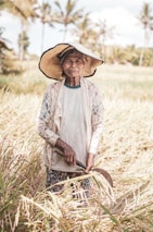 An elderly person stands in a rice field holding a sickle, wearing a wide-brimmed straw hat, a worn-out lace jacket over a plain shirt, and floral patterned pants. The field is filled with tall, golden rice plants swaying gently. Palm trees are visible in the blurred background against a blue sky.