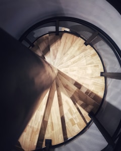 A top-down view of a spiral staircase with wooden steps and a central dark pole. The staircase has a circular layout, with shadows casting intricate patterns on the steps. The surrounding railing appears to have a net-like texture.