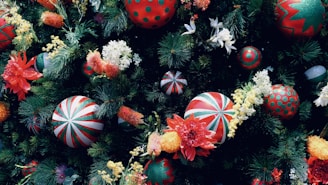 A vibrant arrangement of Christmas ornaments and flowers amidst lush green pine branches. The ornaments feature red and green patterns with stripes and dots, while the flowers include red poinsettias and white lilies, adding a festive touch.