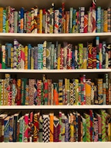 A bookshelf filled with books covered in vibrant, multicolored patterns. The patterns are diverse, featuring geometric shapes, swirling designs, and intricate motifs. The use of bright colors like red, blue, green, orange, and more gives each book a unique, artistic appearance.