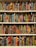 A bookshelf filled with books covered in vibrant, multicolored patterns. The patterns are diverse, featuring geometric shapes, swirling designs, and intricate motifs. The use of bright colors like red, blue, green, orange, and more gives each book a unique, artistic appearance.
