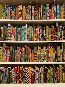 A bookshelf filled with books covered in vibrant, multicolored patterns. The patterns are diverse, featuring geometric shapes, swirling designs, and intricate motifs. The use of bright colors like red, blue, green, orange, and more gives each book a unique, artistic appearance.