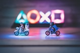 Two small toy figures resembling iconic movie characters ride motorcycles on a reflective surface. Illuminated gaming console symbols in vibrant colors are blurred in the background, creating a playful yet dynamic atmosphere in the dimly lit setting.