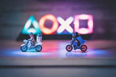 Two small toy figures resembling iconic movie characters ride motorcycles on a reflective surface. Illuminated gaming console symbols in vibrant colors are blurred in the background, creating a playful yet dynamic atmosphere in the dimly lit setting.