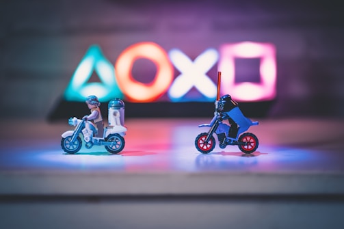 Two small toy figures resembling iconic movie characters ride motorcycles on a reflective surface. Illuminated gaming console symbols in vibrant colors are blurred in the background, creating a playful yet dynamic atmosphere in the dimly lit setting.