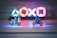 Two small toy figures resembling iconic movie characters ride motorcycles on a reflective surface. Illuminated gaming console symbols in vibrant colors are blurred in the background, creating a playful yet dynamic atmosphere in the dimly lit setting.