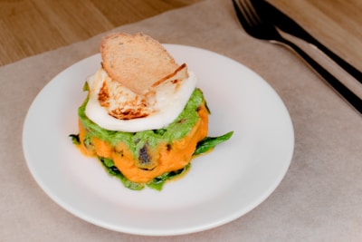A gourmet dish is elegantly presented on a white plate. It features a stack of colorful layers with an orange base, topped with green vegetables and a creamy white sauce. A piece of toasted bread rests on top. The dish is arranged on a light-colored table, accompanied by a fork and knife placed on the right side.