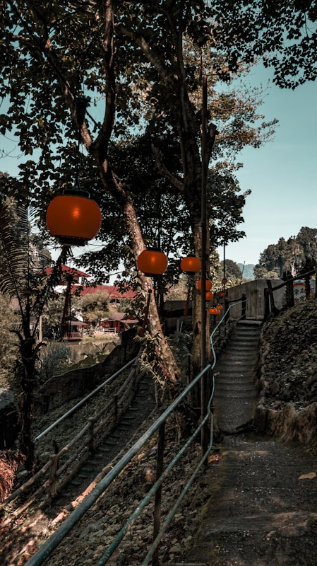 A serene pathway surrounded by lush trees and foliage, adorned with glowing orange lanterns hanging from the branches. The path features a set of stone stairs and is bordered by a rustic wooden railing. In the distance, traditional buildings with red roofs and mountains can be seen, creating a peaceful and tranquil atmosphere.