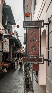 A narrow urban alleyway with signs in multiple languages, including Chinese characters and English text, reading 'Concubine Lane' and 'Since 1908.' The pathway is paved with stone tiles and flanked by small shops and cafes. Red lanterns hang overhead, creating a traditional and vibrant atmosphere. People are walking along the lane, contributing to a bustling and lively scene.