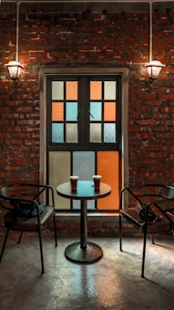 A warm, inviting setting with a brick wall backdrop, illuminated by two vintage wall sconces. A small table with two cups of coffee sits between two empty chairs, facing a window composed of colorful frosted glass panes.