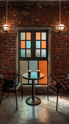 A warm, inviting setting with a brick wall backdrop, illuminated by two vintage wall sconces. A small table with two cups of coffee sits between two empty chairs, facing a window composed of colorful frosted glass panes.