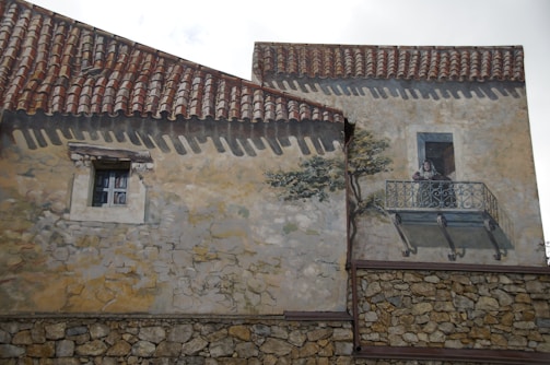 A detailed mural on a building wall mimics the appearance of windows, a tree, and a person standing on a balcony. The painted façade has a realistic look with a textured stone wall, red roof tiles, and the illusion of depth. The person depicted seems to be leaning on the balcony railing.