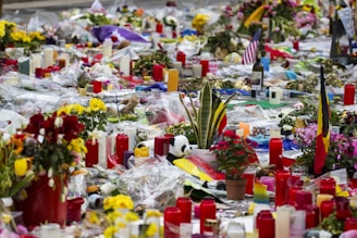 A large collection of flowers, candles, flags, and various memorial items are arranged on the ground in a tribute. Red candles are predominant, surrounded by bouquets in different colors. Small flags, including an American flag and another unidentified flag, are placed among the offerings. The scene is vibrant yet solemn, indicating a memorial or tribute site.