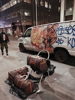 A city street scene at night featuring a white van covered in colorful graffiti parked beside a modern building. Three large designer bags are displayed on the sidewalk, two resting on black trash bags and one on a folding chair. A person walks nearby dressed in dark clothing and camo pants.