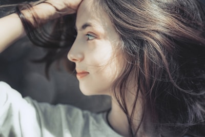A side profile of a woman with long, flowing hair looking thoughtfully into the distance. The lighting highlights her facial features, and she appears to be deep in thought or contemplation.