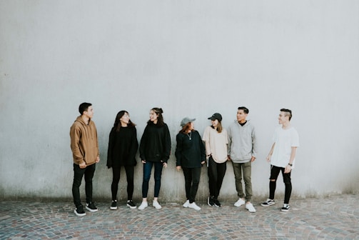 A group of seven young adults stands casually against a plain, light-colored wall. They are dressed in casual clothing, including hoodies, jackets, and jeans, with some wearing hats. The ground is paved with patterned stones. The individuals are interacting with each other, creating a relaxed and friendly atmosphere.