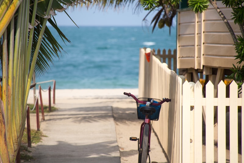 A bicycle leaning against a white picket fence beside a sandy path leading down to a bright turquoise sea