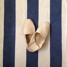 A pair of beige slip-on shoes are placed on a striped rug. The rug features alternating blue and white stripes. The shoes are slightly worn, with visible creases in the fabric.