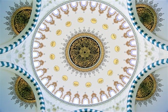 A beautifully intricate dome ceiling featuring detailed geometric and floral patterns. The central circular design includes intricate gold and black motifs surrounded by smaller circular patterns with Arabic calligraphy. The edges of the dome display arch-like structures in a repetitive sequence.