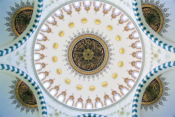 A beautifully intricate dome ceiling featuring detailed geometric and floral patterns. The central circular design includes intricate gold and black motifs surrounded by smaller circular patterns with Arabic calligraphy. The edges of the dome display arch-like structures in a repetitive sequence.