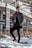 A well-dressed man in a long coat standing confidently in an urban setting.