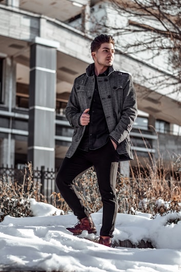 A person stands confidently in a snowy urban setting, dressed in a stylish gray coat, black pants, and brown shoes. The background features a modern building with dark columns and bare trees, suggesting a winter scene.