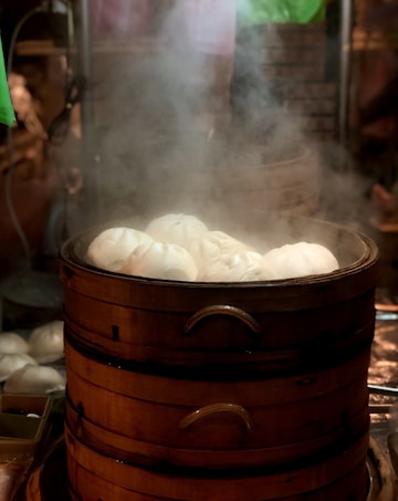A stack of bamboo steamers filled with steaming white buns, emitting a misty vapor. The setting appears to be a kitchen or a street food stall, with a warm and inviting atmosphere.