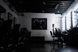 A dimly lit salon with multiple styling chairs positioned along a mirrored wall. The space features exposed wooden beams on the ceiling. A piece of modern artwork hangs on a white brick wall opposite a row of chairs. Vanity mirrors with bright lights are visible on the right, reflecting the furniture and adding a contrast of light.