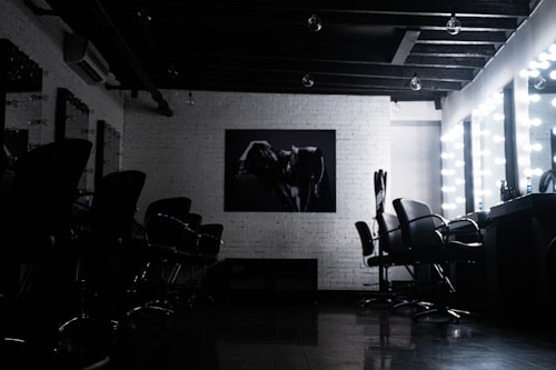 A dimly lit salon with multiple styling chairs positioned along a mirrored wall. The space features exposed wooden beams on the ceiling. A piece of modern artwork hangs on a white brick wall opposite a row of chairs. Vanity mirrors with bright lights are visible on the right, reflecting the furniture and adding a contrast of light.