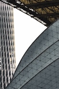 An architectural scene featuring intersecting modern structures with geometric patterns. One structure displays a grid of triangular panes, while another is a cylindrical building with vertical lines. Overhead, a series of interconnected metal beams form a canopy.