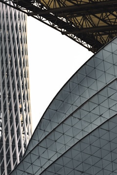 An architectural scene featuring intersecting modern structures with geometric patterns. One structure displays a grid of triangular panes, while another is a cylindrical building with vertical lines. Overhead, a series of interconnected metal beams form a canopy.