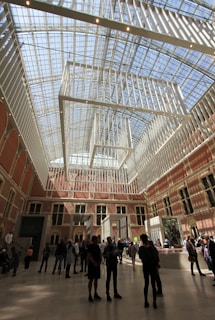 A spacious hall features high ceilings with a modern grid-like framework and large glass windows, allowing ample natural light to flood the room. The interior is adorned with warm-colored brick walls and multiple arched windows. Several groups of people are walking and standing around, engaging in conversation and observing the surroundings. Artistic installations and potted plants are also present.