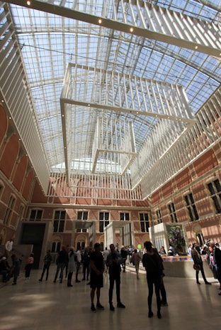A spacious hall features high ceilings with a modern grid-like framework and large glass windows, allowing ample natural light to flood the room. The interior is adorned with warm-colored brick walls and multiple arched windows. Several groups of people are walking and standing around, engaging in conversation and observing the surroundings. Artistic installations and potted plants are also present.