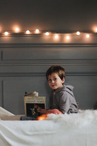 A young child is sitting on a bed holding a book with the title 'Derniers Nomades Grand Nord'. The room is softly lit by a string of warm fairy lights on the wall.