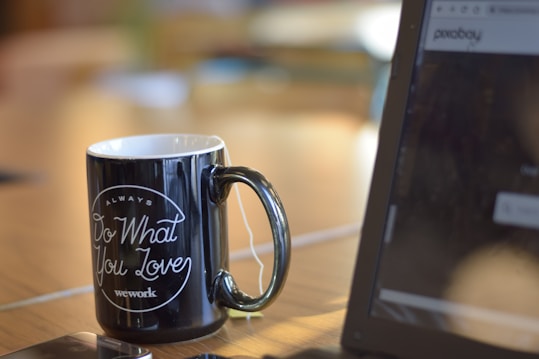 A black mug with the phrase 'Always Do What You Love' printed in white sits on a wooden desk. Next to it, there's an open laptop displaying a website. The mug appears to have a teabag inside, indicated by the string hanging over the side. Soft lighting creates a warm and inviting atmosphere.