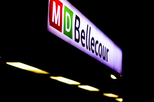An illuminated sign displaying the word 'Bellecour' with 'M' and 'D' in red and green boxes. The sign is against a dark background, creating a strong contrast with the bright letters.
