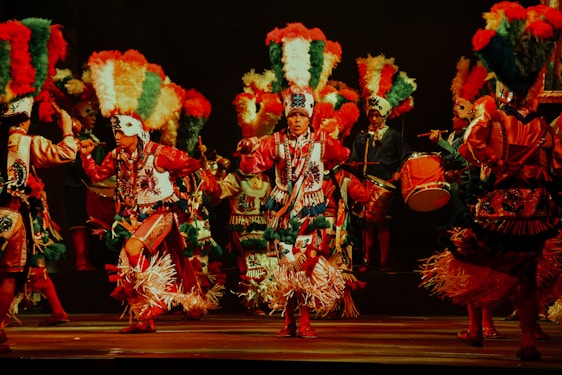 A group of performers dressed in vibrant, festive costumes with large, colorful feathered headdresses. They appear to be engaged in a traditional dance, with some playing musical instruments like drums. The scene is illuminated by stage lighting, enhancing the vividness of their attire.