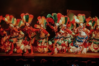 A group of dancers adorned in traditional costumes and vibrant headdresses perform on stage. The attire is richly decorated with feathers, beads, and textiles in vivid colors such as red, green, blue, and white. The dancers appear to be in motion, capturing the energy and dynamism of the performance.