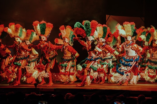 A group of dancers adorned in traditional costumes and vibrant headdresses perform on stage. The attire is richly decorated with feathers, beads, and textiles in vivid colors such as red, green, blue, and white. The dancers appear to be in motion, capturing the energy and dynamism of the performance.